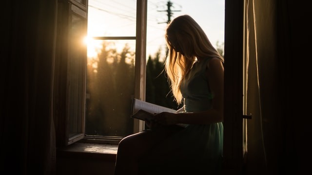 Woman reading book while sitting on windowsill